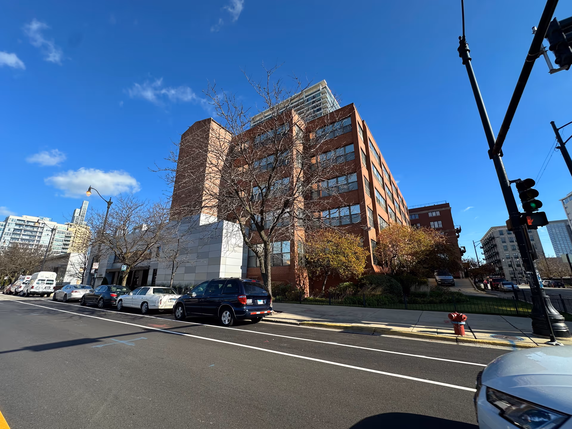Exterior view of a multi-story brick building with large windows on a sunny day, with several parked cars along the street and leafless trees in front of the building.