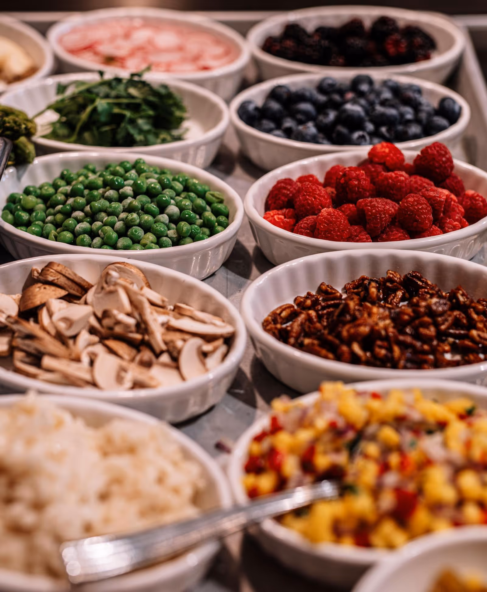Close-up of white bowls on a buffet filled with assorted salad ingredients and toppings including peas, raspberries, blueberries, mushrooms, nuts and rice.