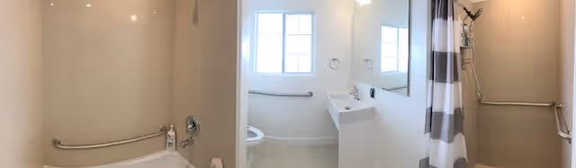Panoramic view of a clean, accessible bathroom featuring a bathtub with grab bars and a shower curtain on the left, a toilet positioned under a window in the center, and a sink with a mirror and towel rings on the right.
