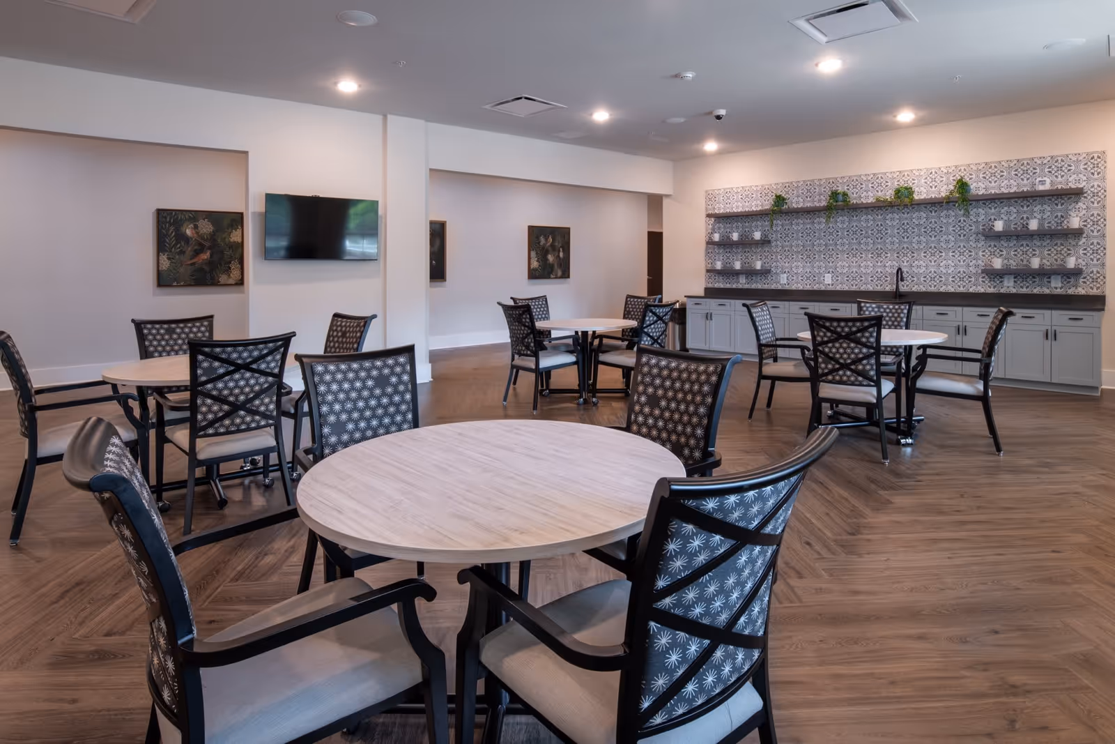 A spacious dining area with several round tables and chairs arranged neatly. The room features a patterned backsplash with shelves holding small plants and decorative items. There is a wall-mounted TV and framed artwork on the walls. The floor has a wood-like finish and the ceiling has recessed lighting.
