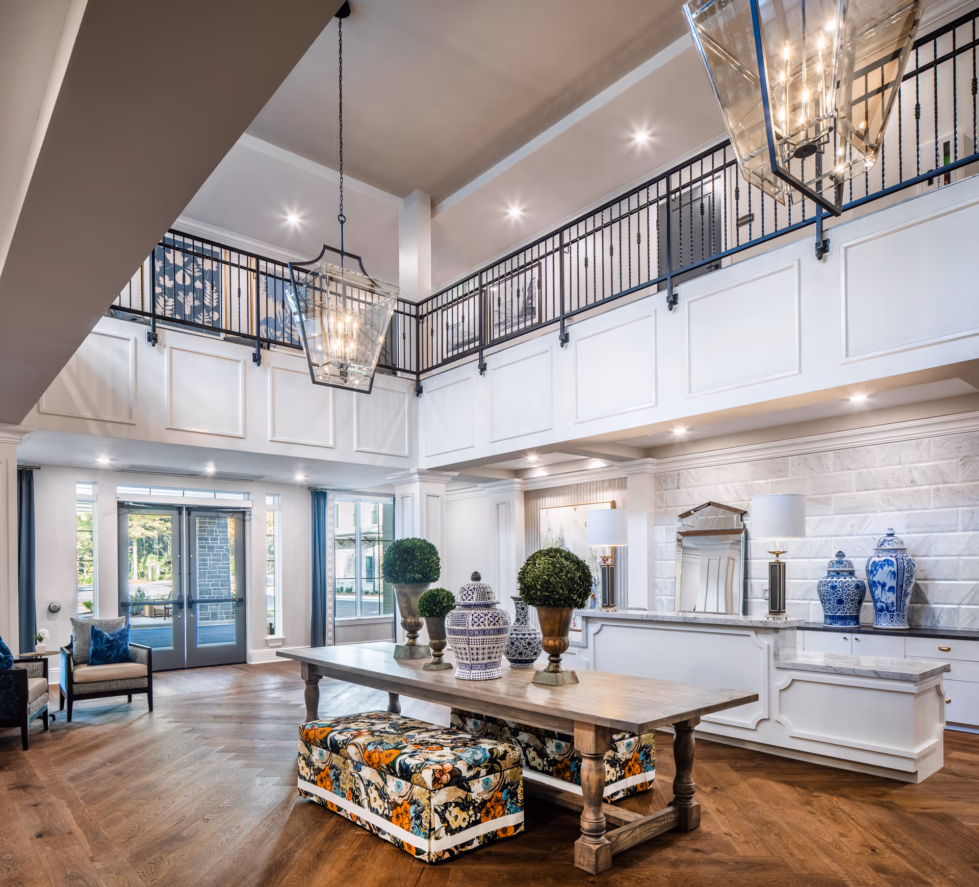 Spacious and elegant senior living facility lobby with high ceilings, large hanging lantern-style light fixtures, a wooden table decorated with potted plants and blue-and-white ceramic vases, floral patterned benches underneath the table, and seating area with chairs near large windows and glass doors allowing natural light.