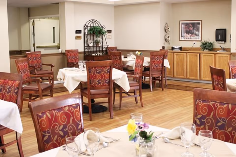 Dining room in Pomerelle Place Senior Living with several tables covered with white tablecloths, set with glassware, silverware, and napkins. Chairs with red patterned upholstery surround the tables. The room has wooden flooring, beige walls, a sideboard with cabinets, and decorative plants and artwork on the walls.