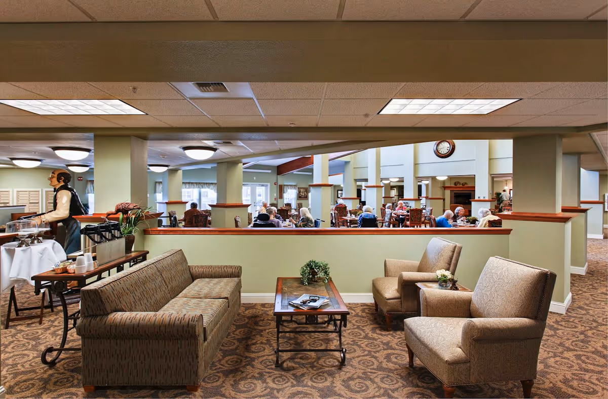 Interior view of a senior living community common area with a seating arrangement including a patterned sofa, two armchairs, and a coffee table with magazines and a small plant. In the background, several elderly residents are seated at dining tables. The room has green walls, carpeted floors, and ceiling lights. A waiter statue is positioned near a table with coffee and cups.