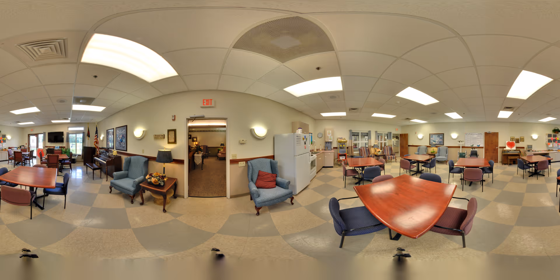 Large communal dining and activity room with multiple tables and chairs, a kitchenette, and lounge seating.