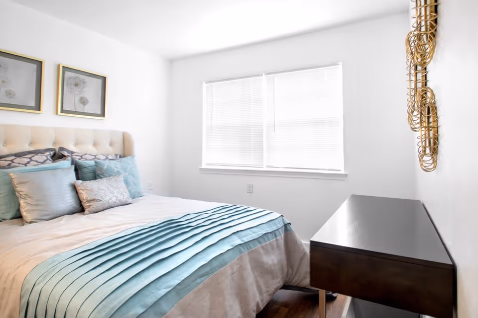 Bright bedroom with a bed dressed in teal-accented bedding, a window with blinds, and a dark dresser.