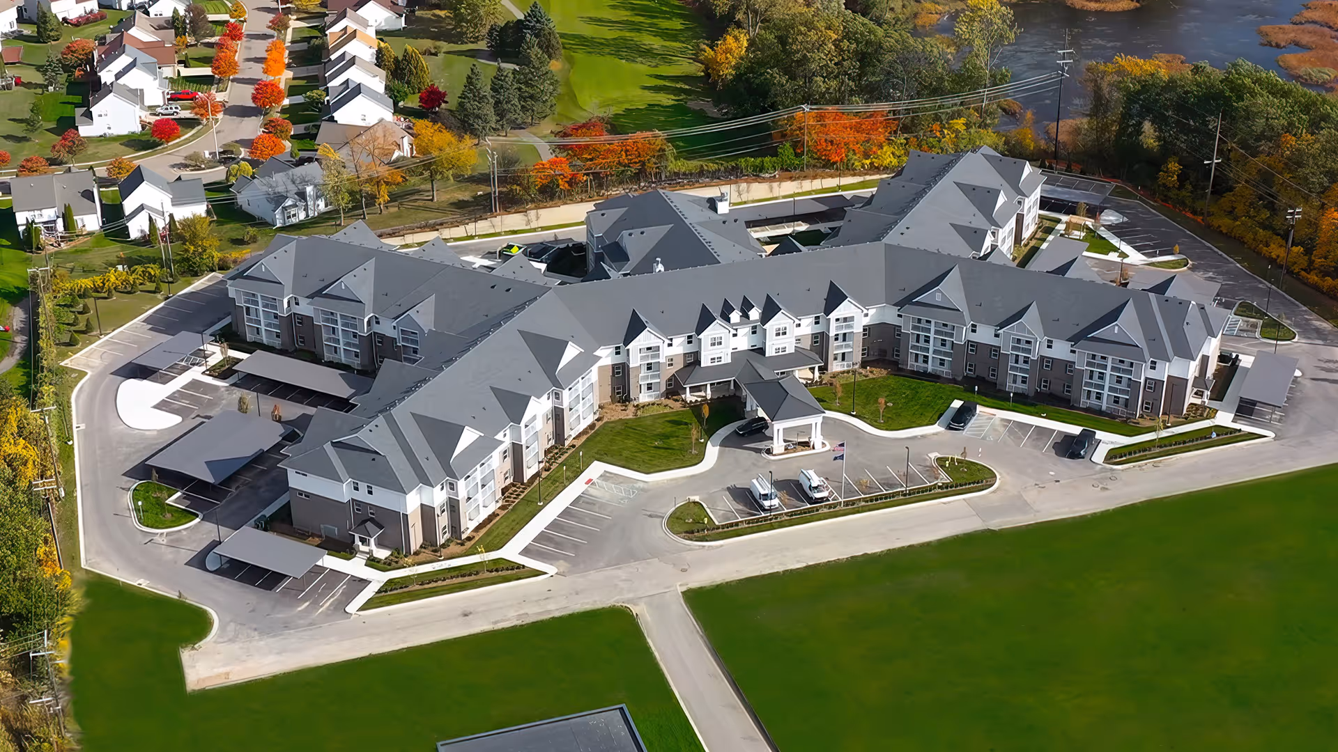 Aerial view of a large senior living facility named American House Village at Bloomfield, featuring a multi-wing building with gray roofs, surrounded by parking lots, green lawns, and nearby residential houses with autumn-colored trees.
