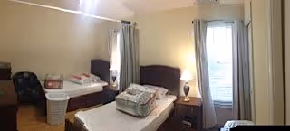 A simple bedroom with two single beds, each with pillows and folded blankets. There is a small bedside table with a lamp between the beds, a laundry basket, a chair, and windows with curtains allowing natural light into the room.