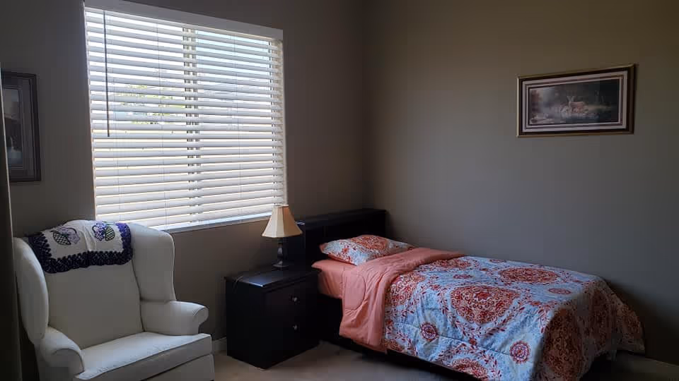 A small bedroom with a single bed covered in a red and white patterned quilt and a matching pillow. Next to the bed is a black nightstand with a small lamp. A white armchair with a decorative throw is positioned near a window with white blinds. A framed picture hangs on the wall above the bed.