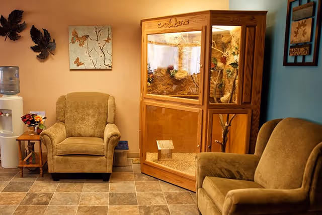 A cozy seating area with two upholstered armchairs, a wooden glass terrarium, a side table with flowers, a water cooler, and wall art on the walls.
