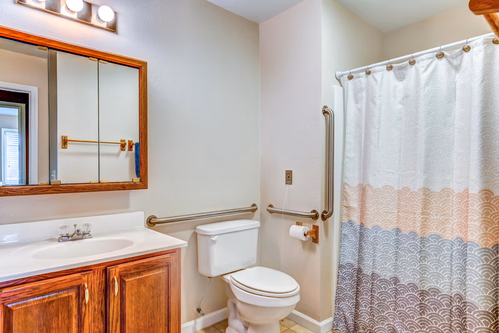 A bathroom with a white toilet, a wooden vanity with a white sink, a large mirror with wooden frame above the sink, and a shower with a patterned curtain in shades of white, orange, blue, and brown. The bathroom has beige walls and grab bars installed near the toilet and shower for accessibility.