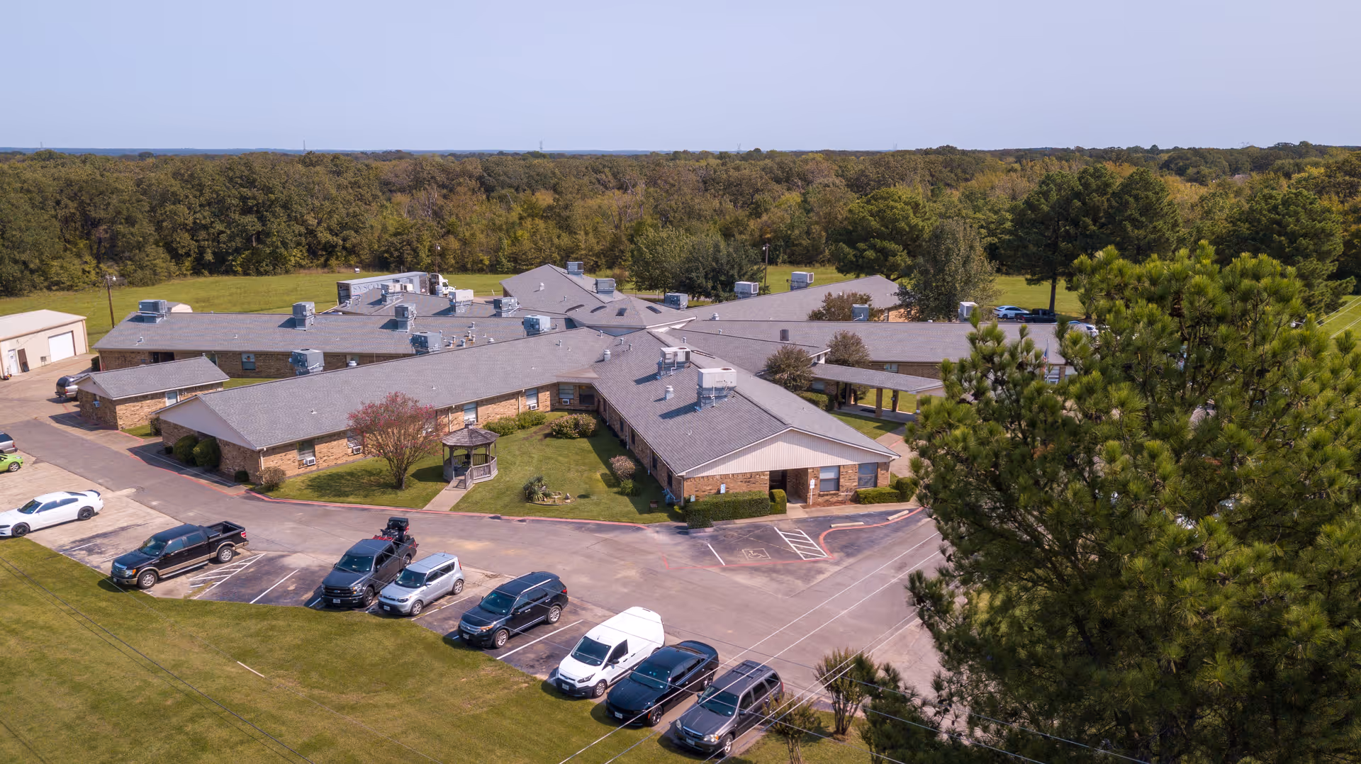 Aerial view of a single-story senior living facility with multiple wings forming a star-like shape, surrounded by green lawns and trees. Several cars are parked in the parking lot in front of the building, and a small gazebo is visible in the courtyard area. The background shows a dense forested area under a clear sky.