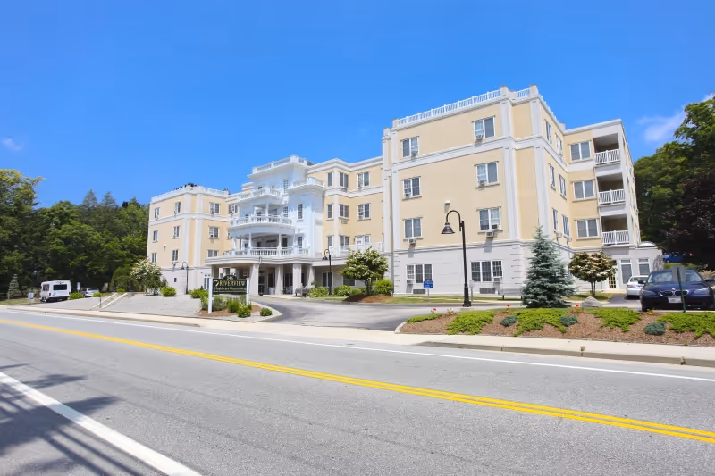 Exterior view of a large, multi-story beige and white senior living facility building with balconies and a covered entrance. The building is surrounded by landscaped greenery and a paved road with yellow dividing lines in front. A sign near the entrance reads 'Riverview Rehabilitation & Healthcare Center.'