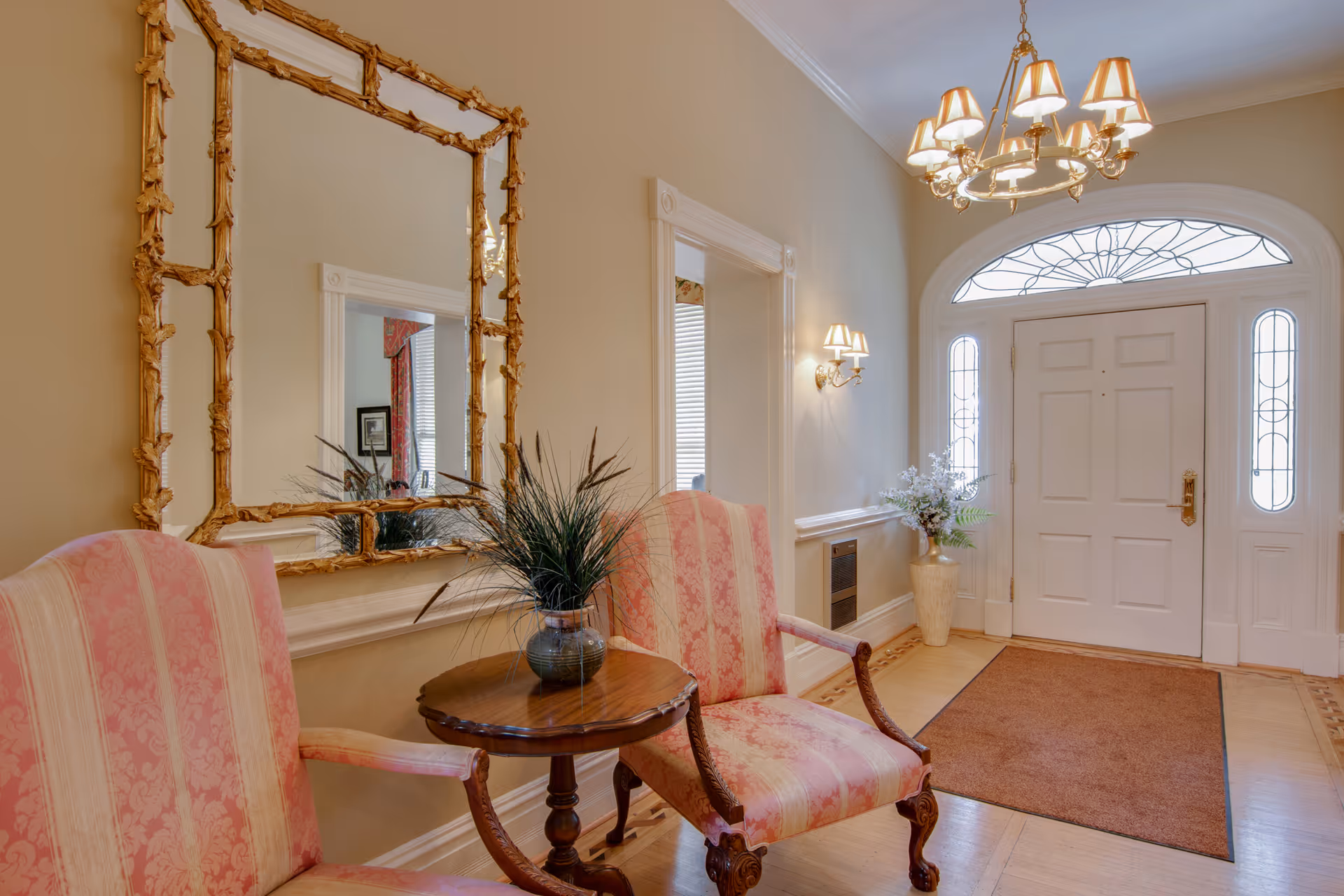 An elegant entryway with a white front door featuring decorative glass panels. Two pink upholstered armchairs with wooden frames flank a small round wooden table holding a vase with green plants. A large ornate gold-framed mirror hangs on the beige wall above the chairs. A chandelier with multiple lampshades hangs from the ceiling, and a wall sconce with two lampshades is mounted on the wall. A brown rug lies on the wooden floor in front of the door.