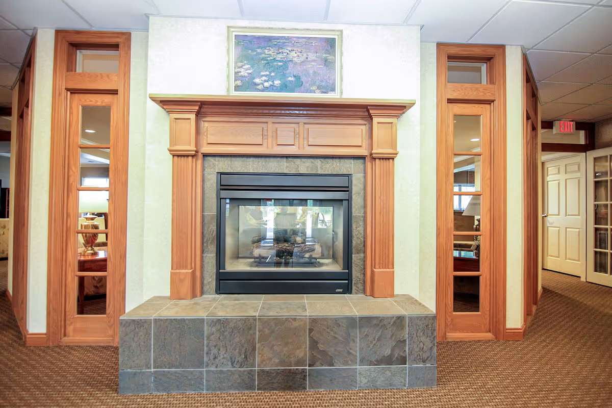 Interior view of a senior living facility featuring a central fireplace with a wooden mantel and tiled base. On either side of the fireplace are tall wooden framed windows with glass panels. Above the mantel hangs a framed painting of water lilies. The room has beige walls, a carpeted floor, and a drop ceiling with fluorescent lighting. Doors and additional rooms are visible in the background.