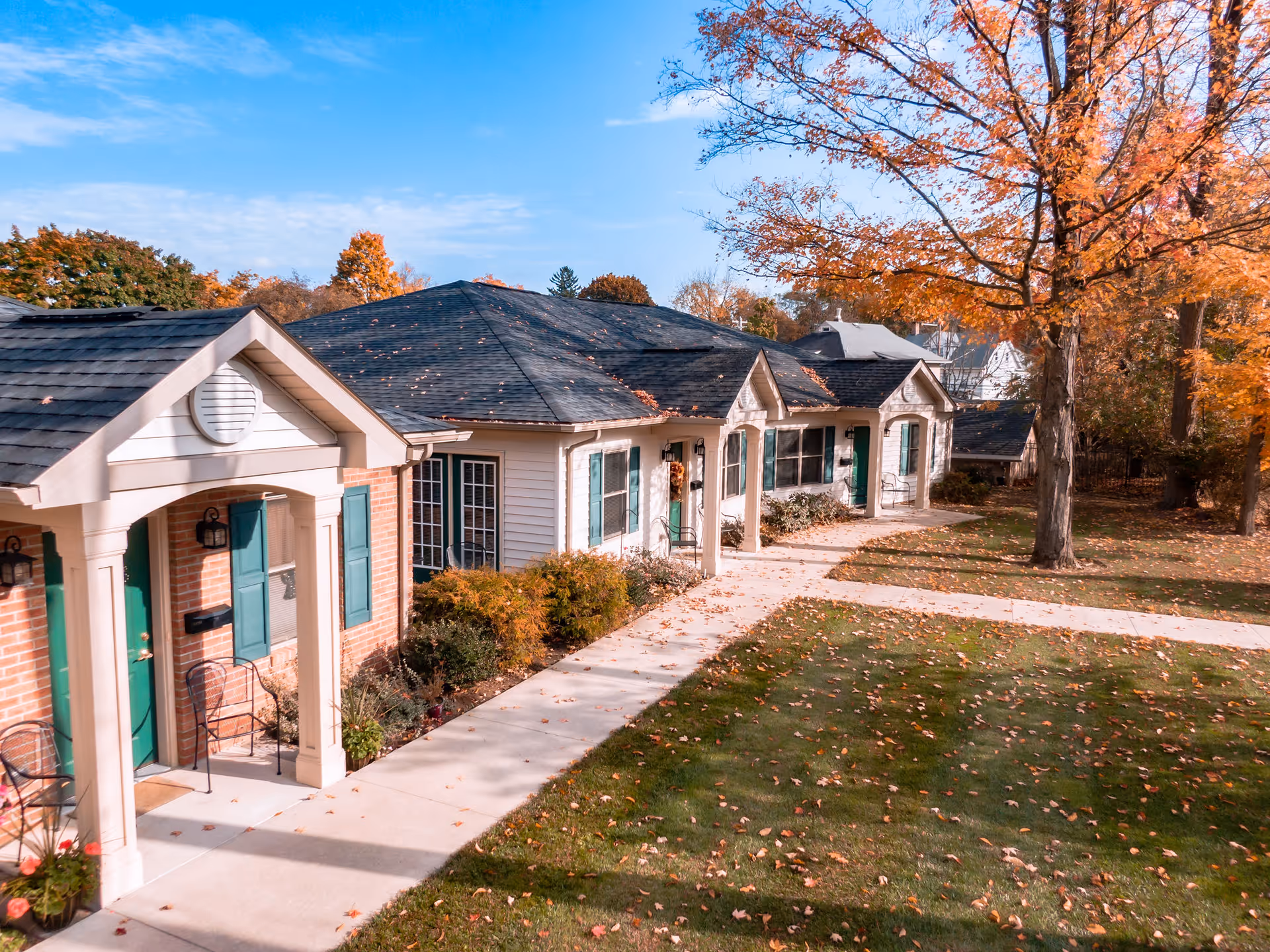 Exterior view of a senior living facility with single-story buildings featuring green doors and windows, surrounded by autumn trees with orange leaves and a well-maintained lawn with a concrete walkway.
