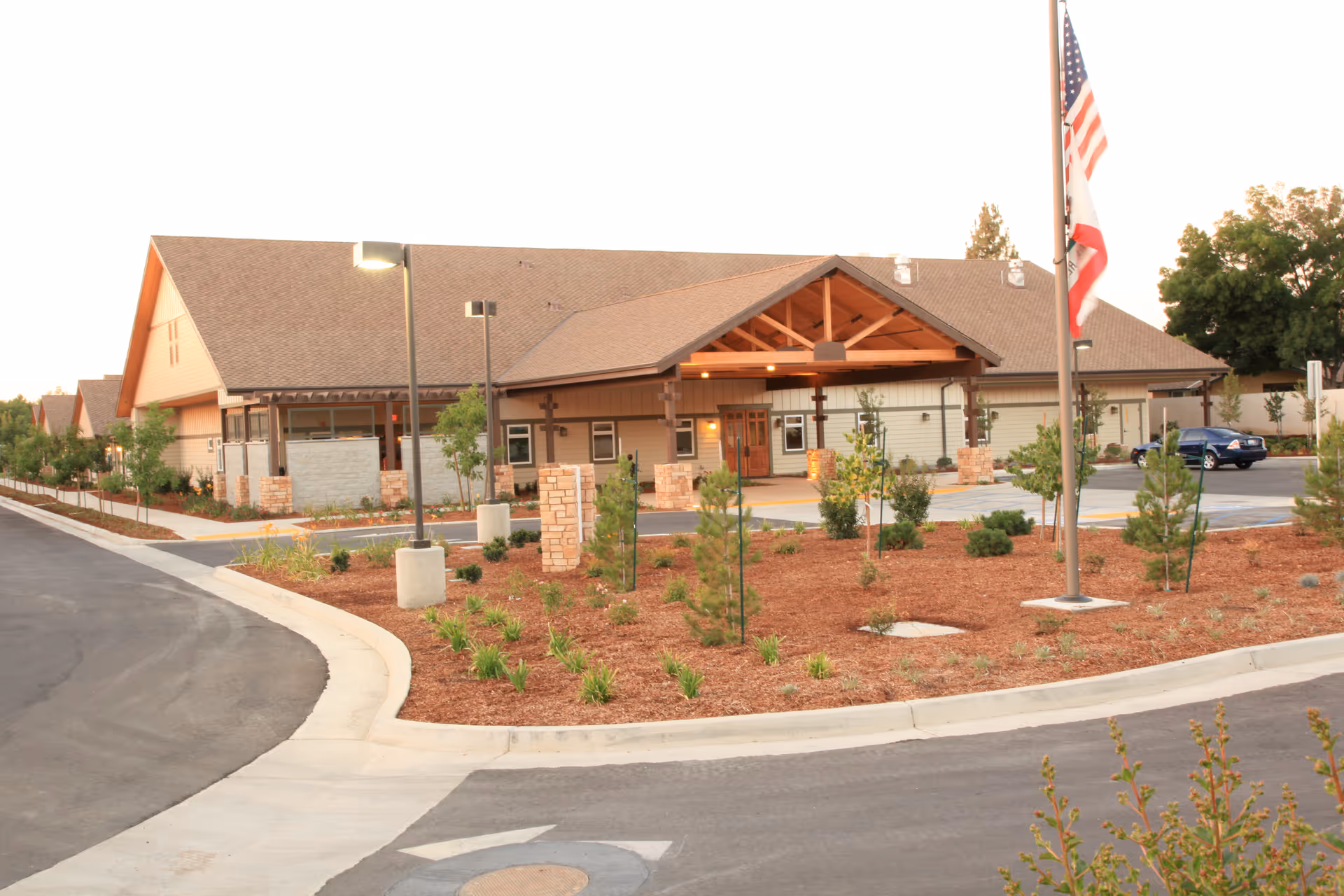 Exterior view of Quail Park Memory Care Residences of Visalia, showing a single-story building with a large covered entrance, landscaped garden with small trees and shrubs, a flagpole with American and California flags, and a parking area with a car.