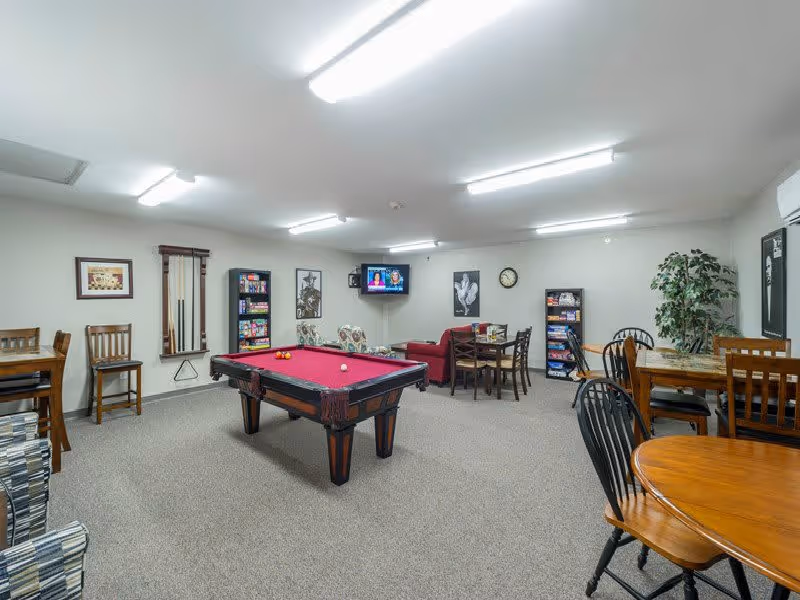 A spacious recreational room with a red pool table in the center, surrounded by various seating options including wooden chairs, a small sofa, and armchairs. The room has bright overhead lighting, a wall-mounted TV, framed pictures, a clock, and shelves stocked with board games. The walls are light-colored and the floor is carpeted.