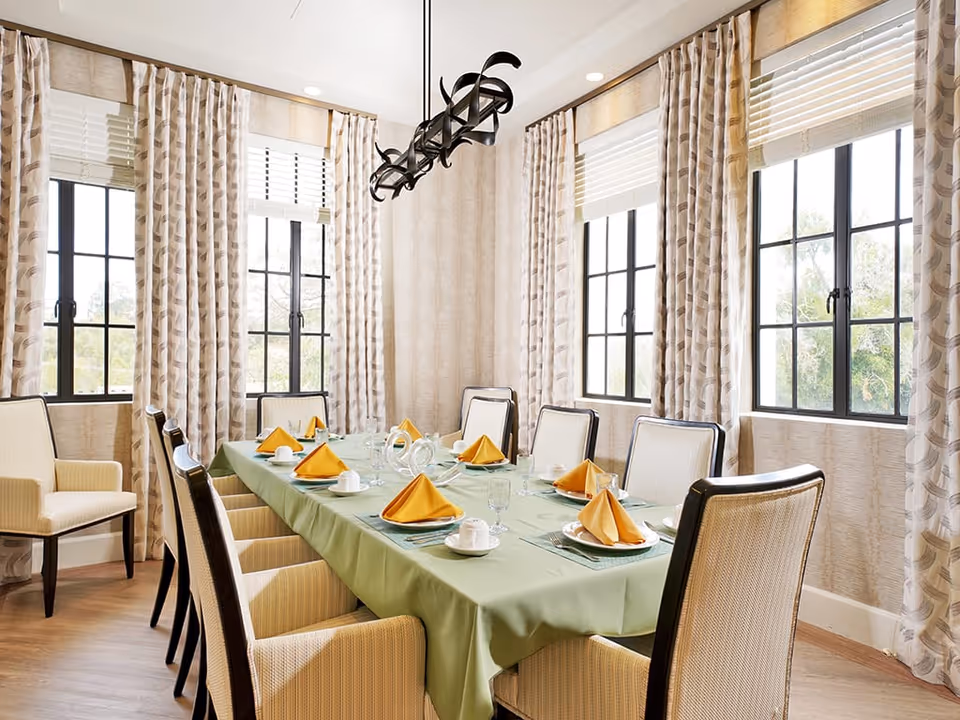 A bright dining room with a long rectangular table covered with a green tablecloth. The table is set with plates, glasses, cups, and yellow folded napkins. The room has large windows with patterned curtains and light-colored walls. Several cushioned chairs with armrests surround the table.