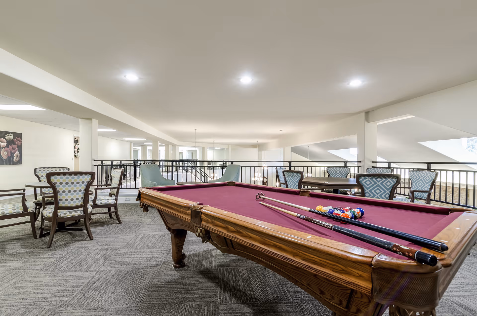 A spacious indoor recreational area featuring a pool table with a burgundy felt surface and pool cues resting on it. Surrounding the pool table are several seating arrangements including patterned upholstered chairs and small tables. The area is well-lit with ceiling lights and has a railing overlooking a lower level.