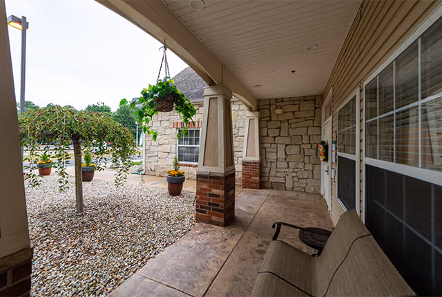 Covered outdoor patio area with a bench, hanging plant, and potted plants. The patio has stone and brick pillars and overlooks a landscaped area with gravel and small trees.