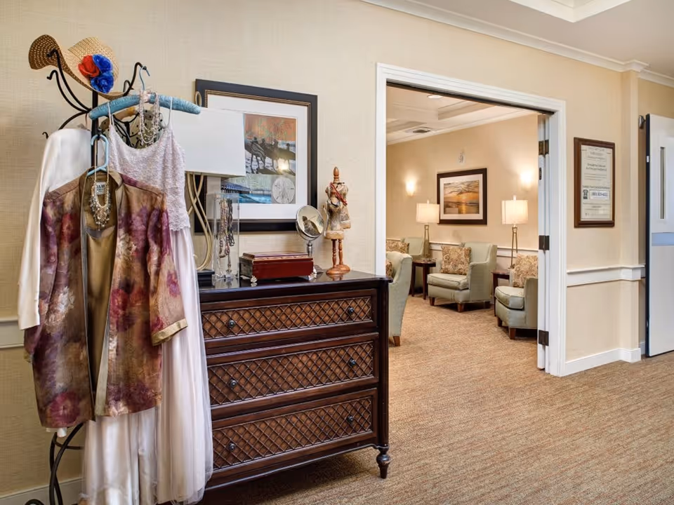 Interior view of a senior living facility hallway with a decorative coat rack holding clothes and accessories next to a dark wooden chest of drawers. In the background, an open doorway reveals a cozy sitting area with armchairs, side tables, lamps, and framed artwork on the walls.