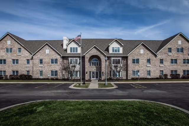 Front exterior view of a large brick senior living facility building with multiple windows, a central entrance with an archway, an American flag on a flagpole, and a paved driveway with parking spaces in front.
