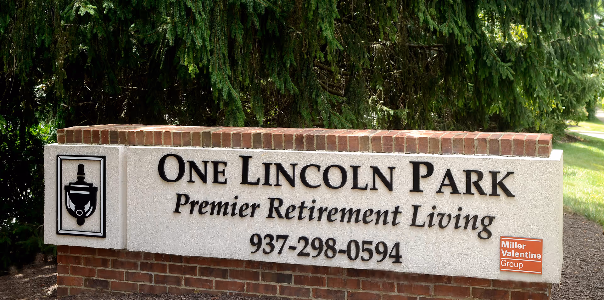 A large outdoor sign for One Lincoln Park Premier Retirement Living, featuring the phone number 937-298-0594. The sign is made of brick and stucco with black lettering, and there is a small red plaque on the bottom right corner that reads Miller Valentine Group. The background includes green trees and grass.