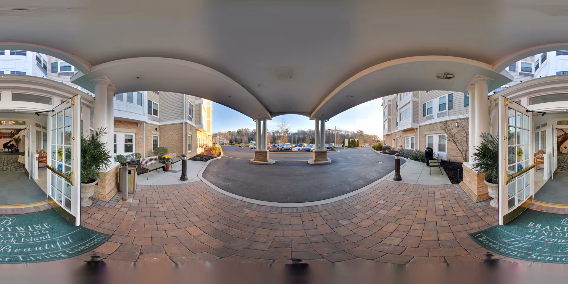 Covered entrance area of Brandywine Fenwick Island by Monarch senior living facility with open glass doors leading inside, brick-paved walkway, benches, potted plants, and a parking lot in the background under a clear sky.