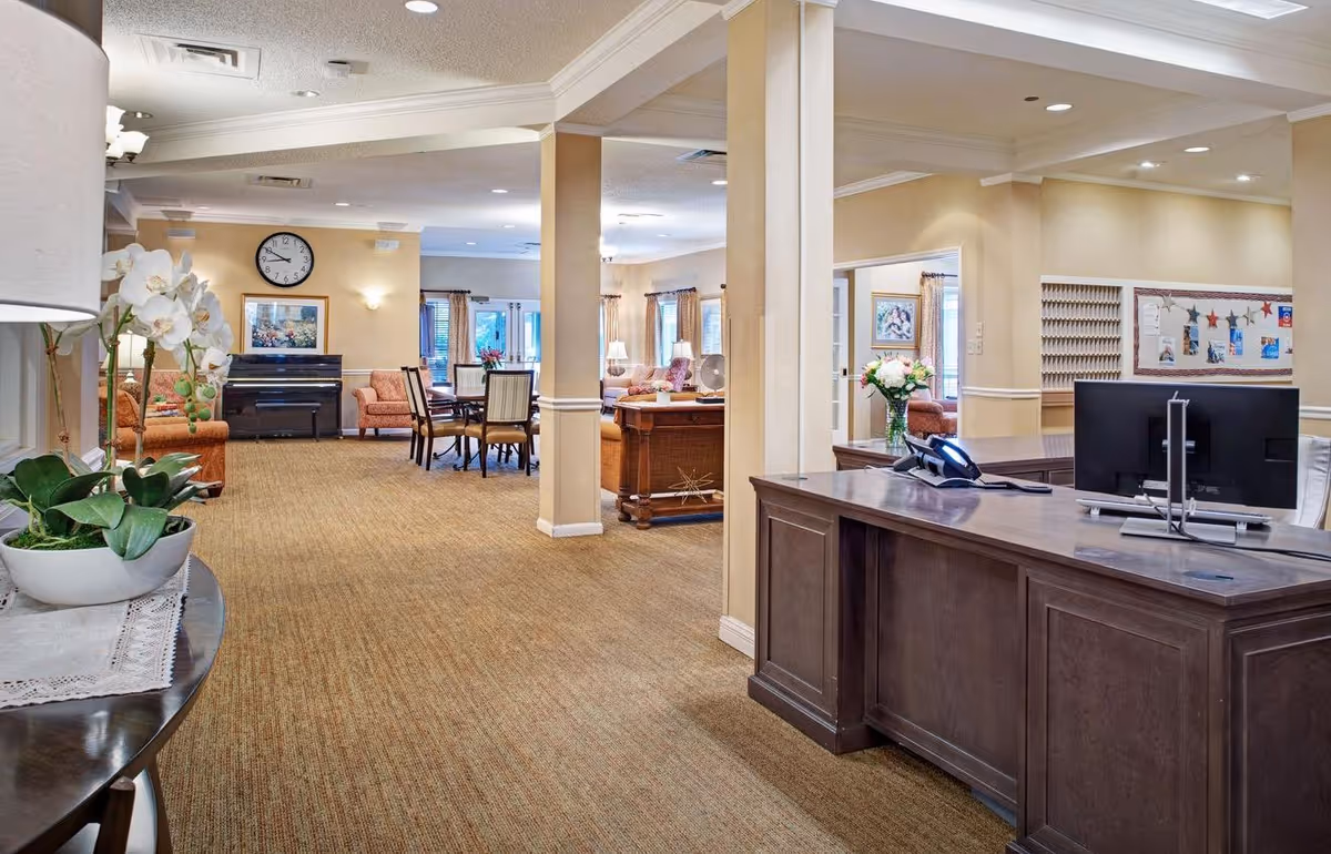 Interior view of a senior living facility lobby and common area with a reception desk on the right, a computer and phone on the desk, a vase with flowers, seating areas with chairs and sofas, a piano against the far wall, a clock above the piano, and soft lighting throughout.