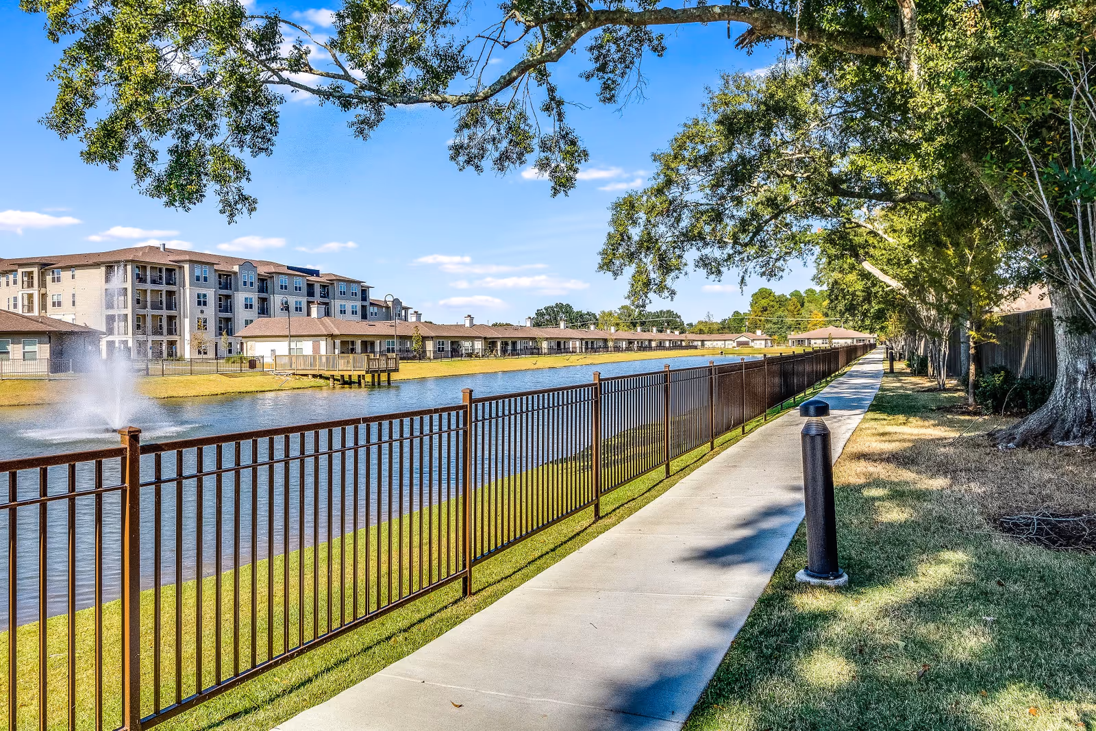 A paved walkway runs alongside a fenced pond with a water fountain, surrounded by green grass and large trees. In the background, there are multi-story residential buildings and single-story structures under a blue sky with scattered clouds.