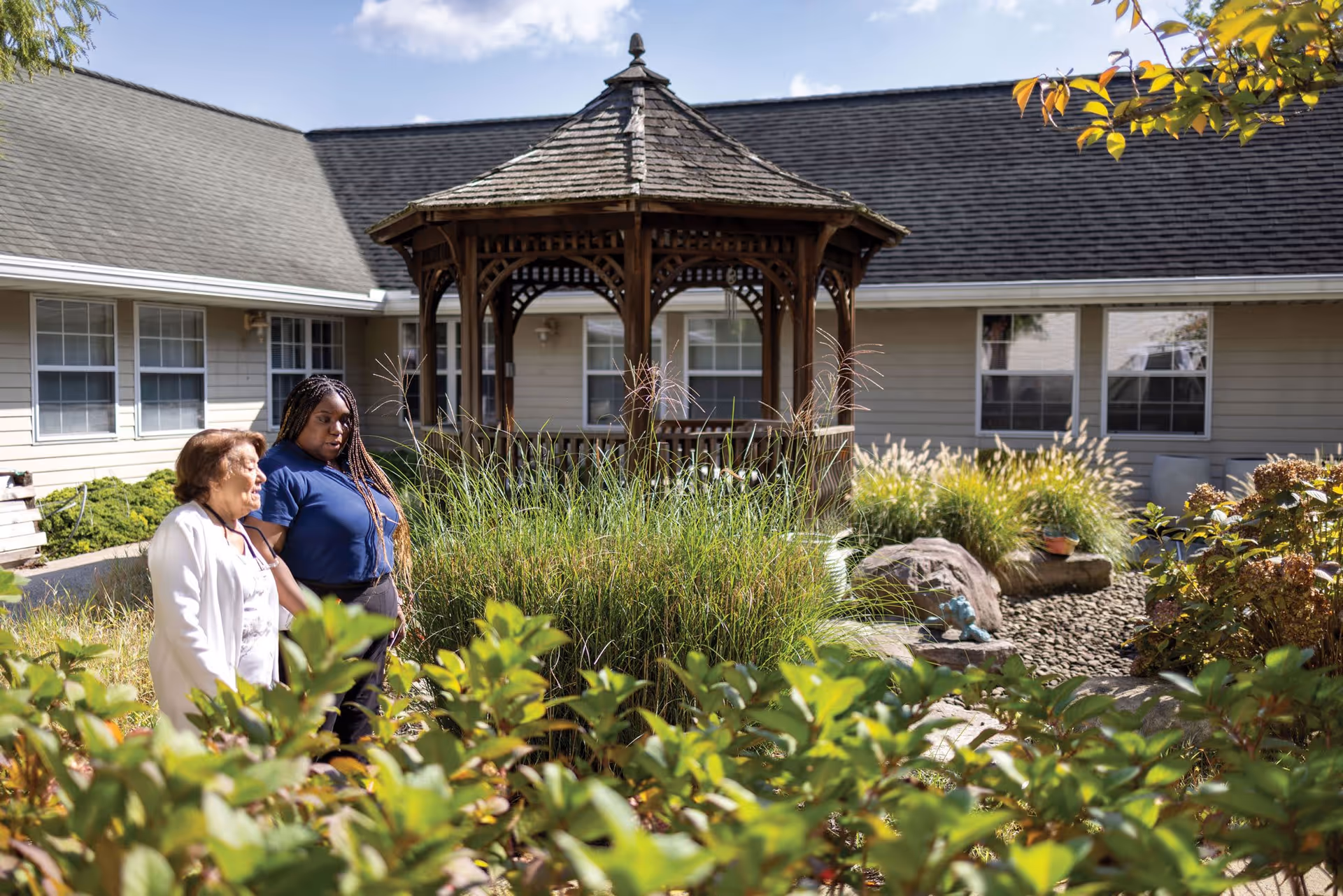 Two women walking together in a garden area with green plants and bushes. In the background, there is a wooden gazebo and a building with multiple windows under a partly cloudy sky.