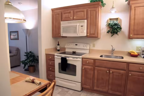 A kitchen with wooden cabinets, a white stove with an oven, a microwave above the stove, a sink with a faucet, and a countertop with a bowl of fruit. There is a dining table with place settings in the foreground and a glimpse of a living room area with a couch and a floor lamp in the background.