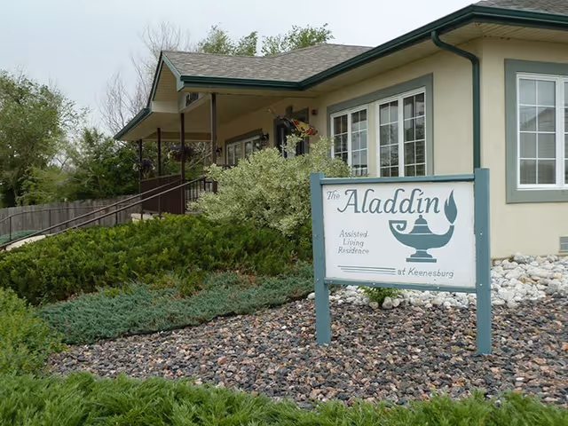 Exterior view of The Aladdin Assisted Living Residence building with a sign in front displaying the facility's name. The building has beige walls, green trim, and multiple windows. There are shrubs, bushes, and rocks in the landscaped area around the sign.