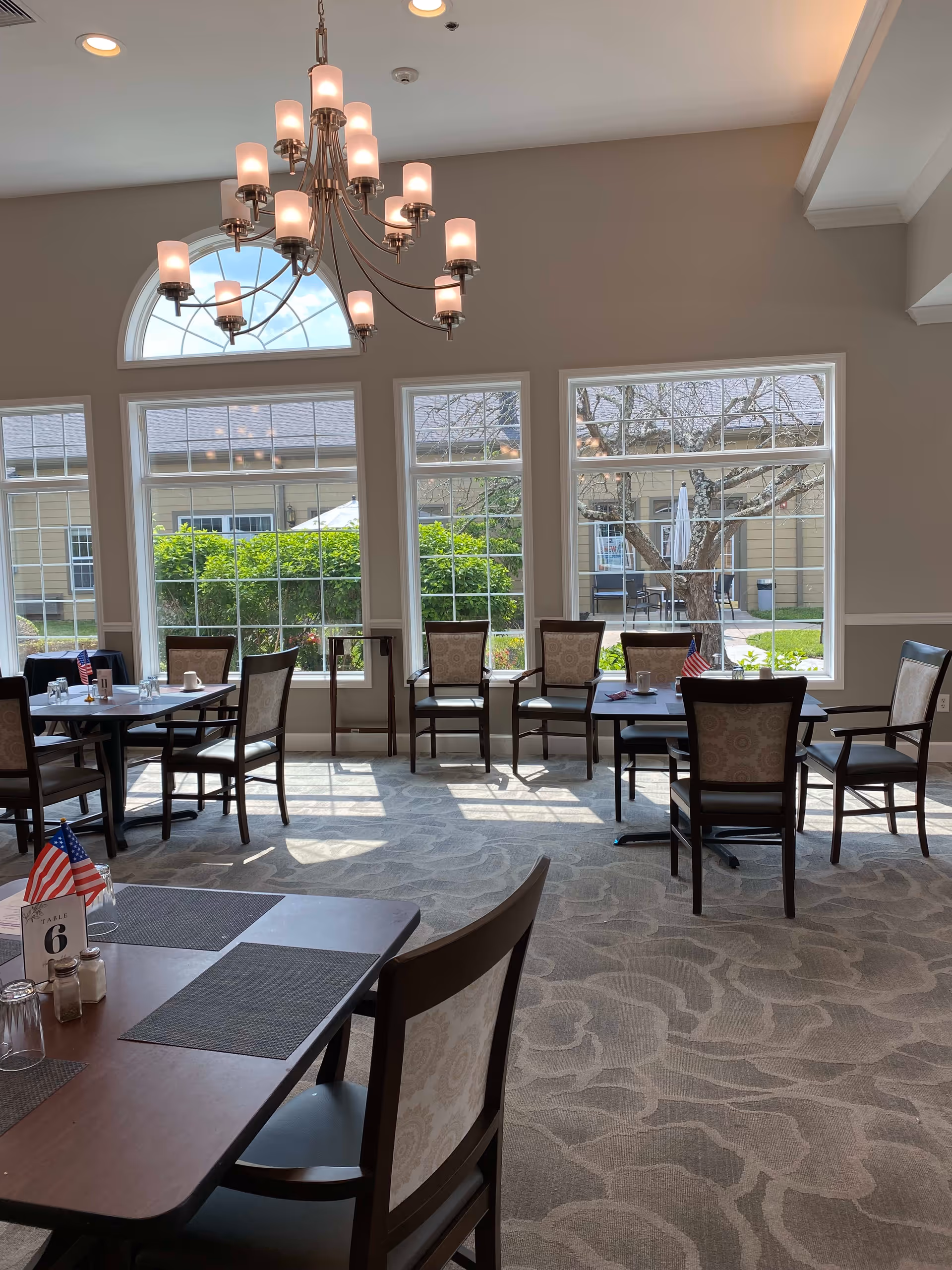 A bright dining room with several tables and chairs arranged neatly. Large windows allow natural light to fill the room, showing greenery and outdoor seating outside. A chandelier with multiple lights hangs from the ceiling.