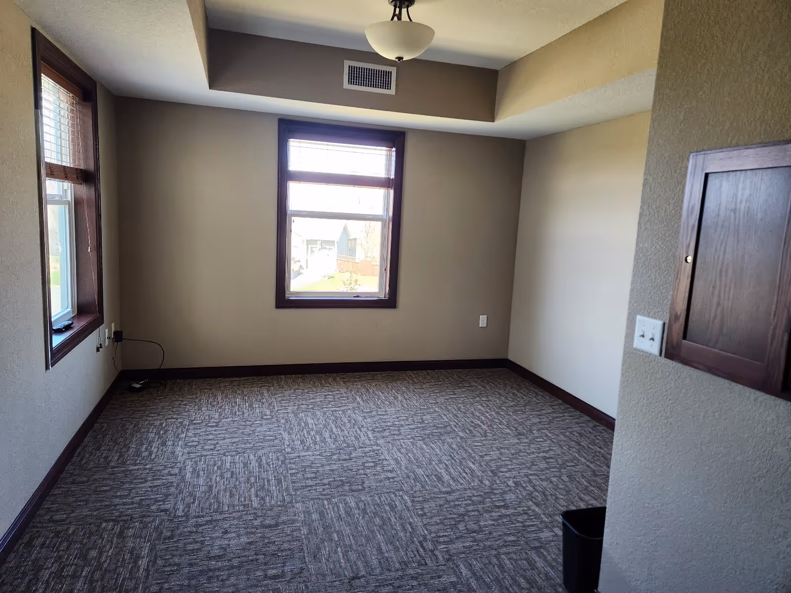 Empty room with beige walls, two windows with wooden frames, a ceiling light fixture, carpeted floor with a patterned design, a small wooden cabinet on the right wall, and electrical outlets visible.