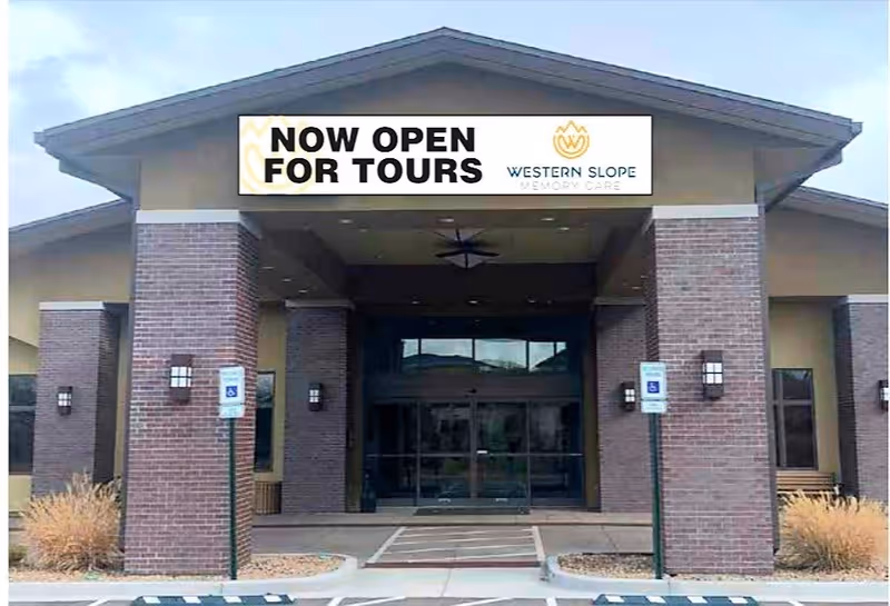 Front entrance of Western Slope Memory Care with a covered drive-up, brick columns and a banner reading "NOW OPEN FOR TOURS".