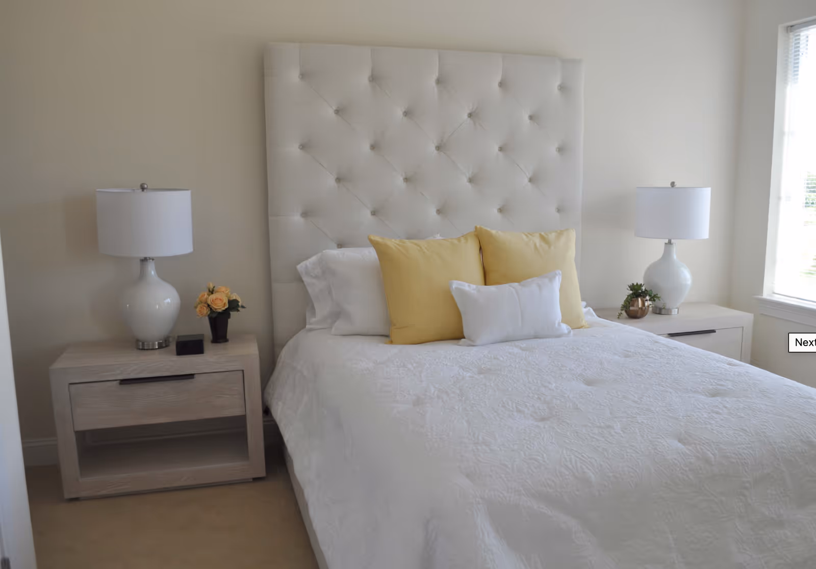 Bright bedroom with a tall tufted white headboard, white quilted bedding and yellow pillows, flanked by matching nightstands with lamps and small decor.