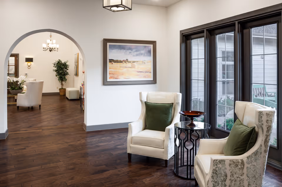 A cozy seating area with two white armchairs featuring green cushions, separated by a small round glass-top table with a decorative bowl. Large windows with dark frames allow natural light to brighten the space. In the background, an arched doorway leads to another room with additional seating, a chandelier, wall sconces, and a potted plant. A framed landscape painting hangs on the wall between the seating area and the doorway.