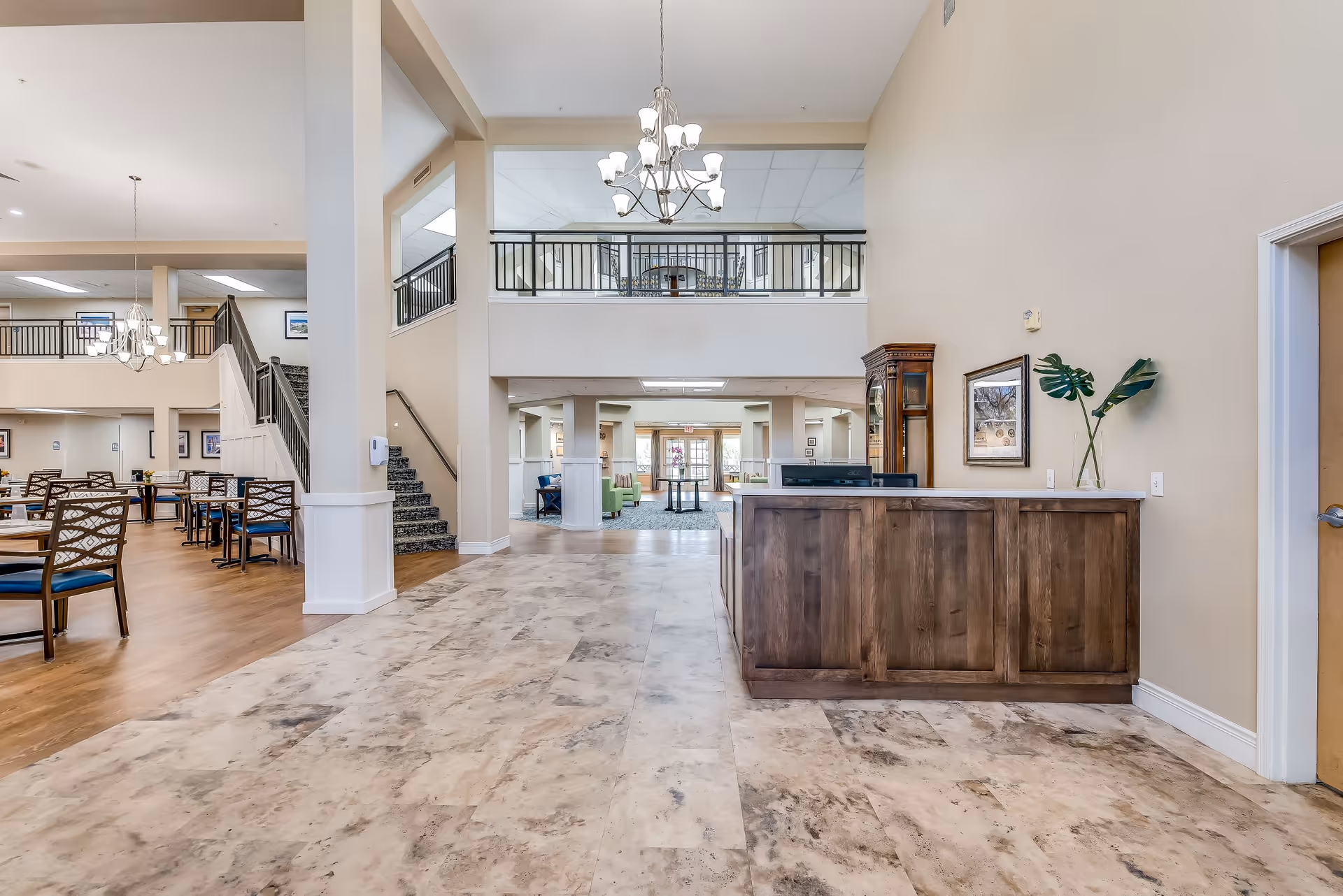 Spacious senior living facility interior with a reception desk on the right, a chandelier hanging from the ceiling, and a dining area with tables and chairs on the left. There is a staircase leading to an upper floor with a black railing, and an open hallway leading further into the building.