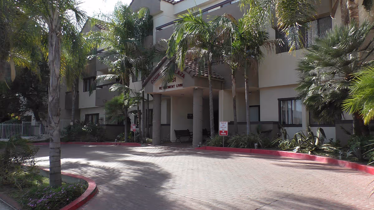 Entrance to Courtyard Plaza retirement living facility with a covered driveway, palm trees, and a bench near the entrance. The building has multiple windows and a tiled roof over the entrance.
