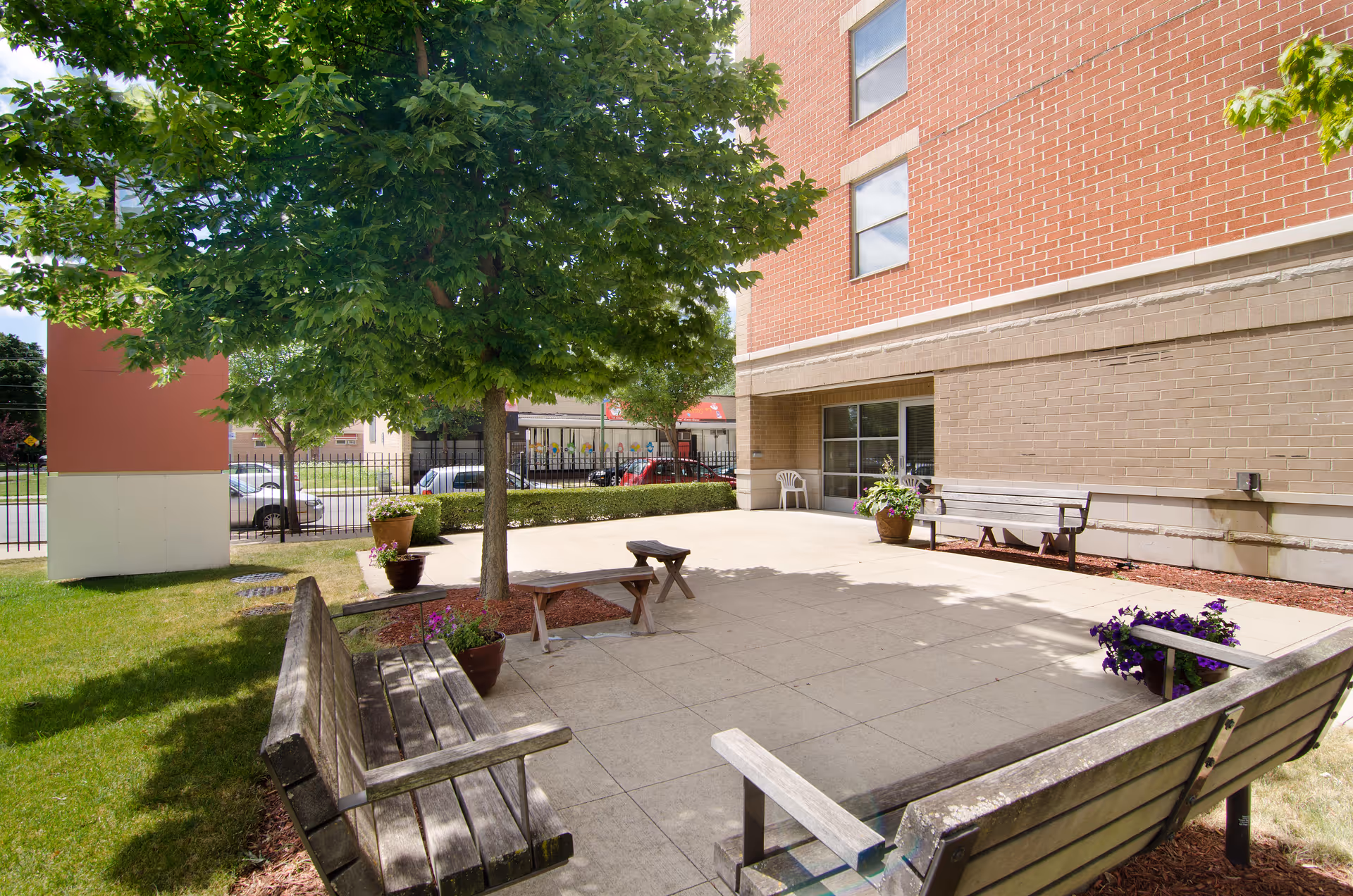 Outdoor patio area at Senior Suites of Washington Heights featuring wooden benches, potted plants, a tree providing shade, and a brick building wall in the background.