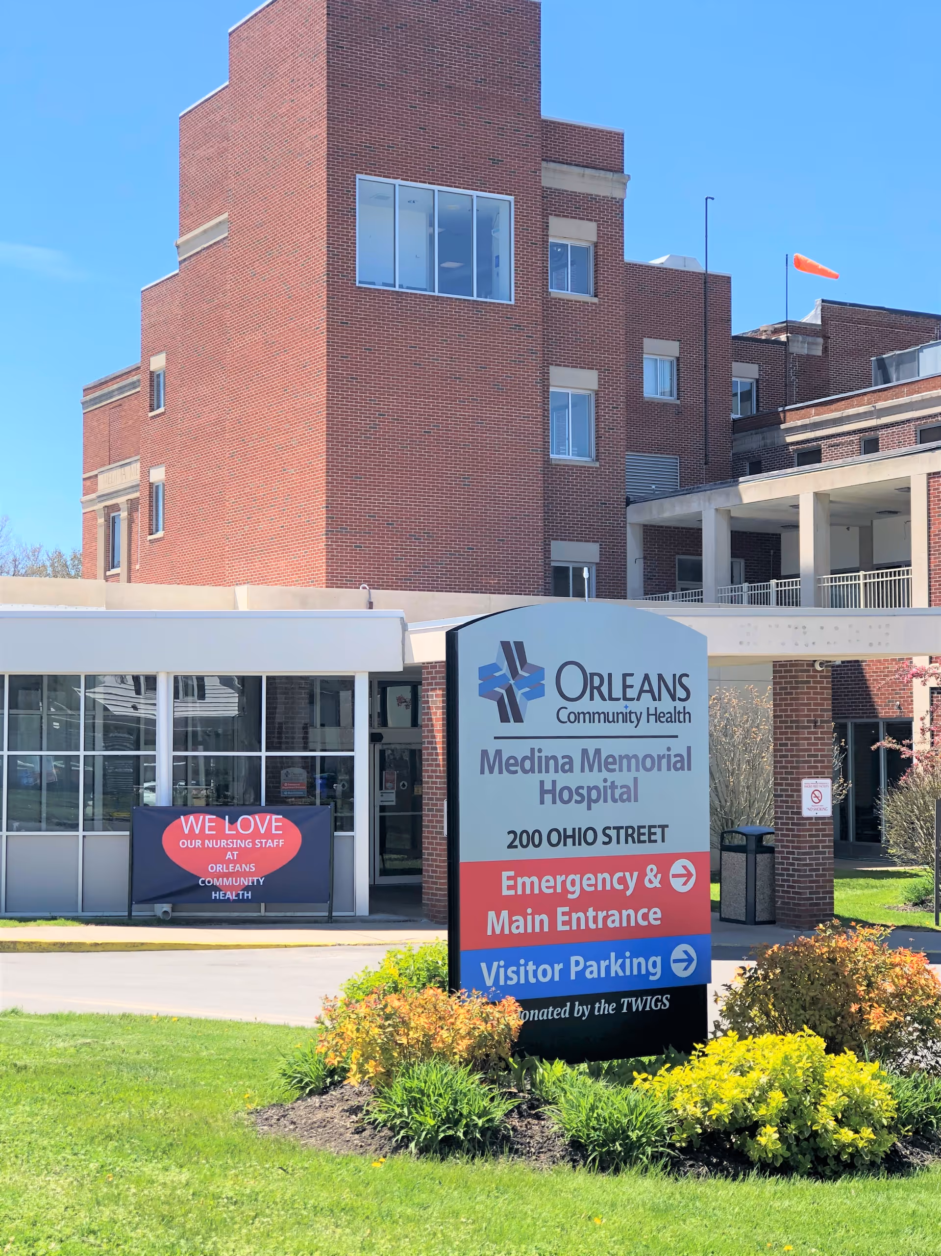 Exterior view of Orleans Community Health Medina Memorial Hospital building with a large sign in front indicating the address 200 Ohio Street, directions for Emergency & Main Entrance, and Visitor Parking. There is a banner on the building that reads 'We love our nursing staff at Orleans Community Health'. The building is made of red brick and there is a clear blue sky above.