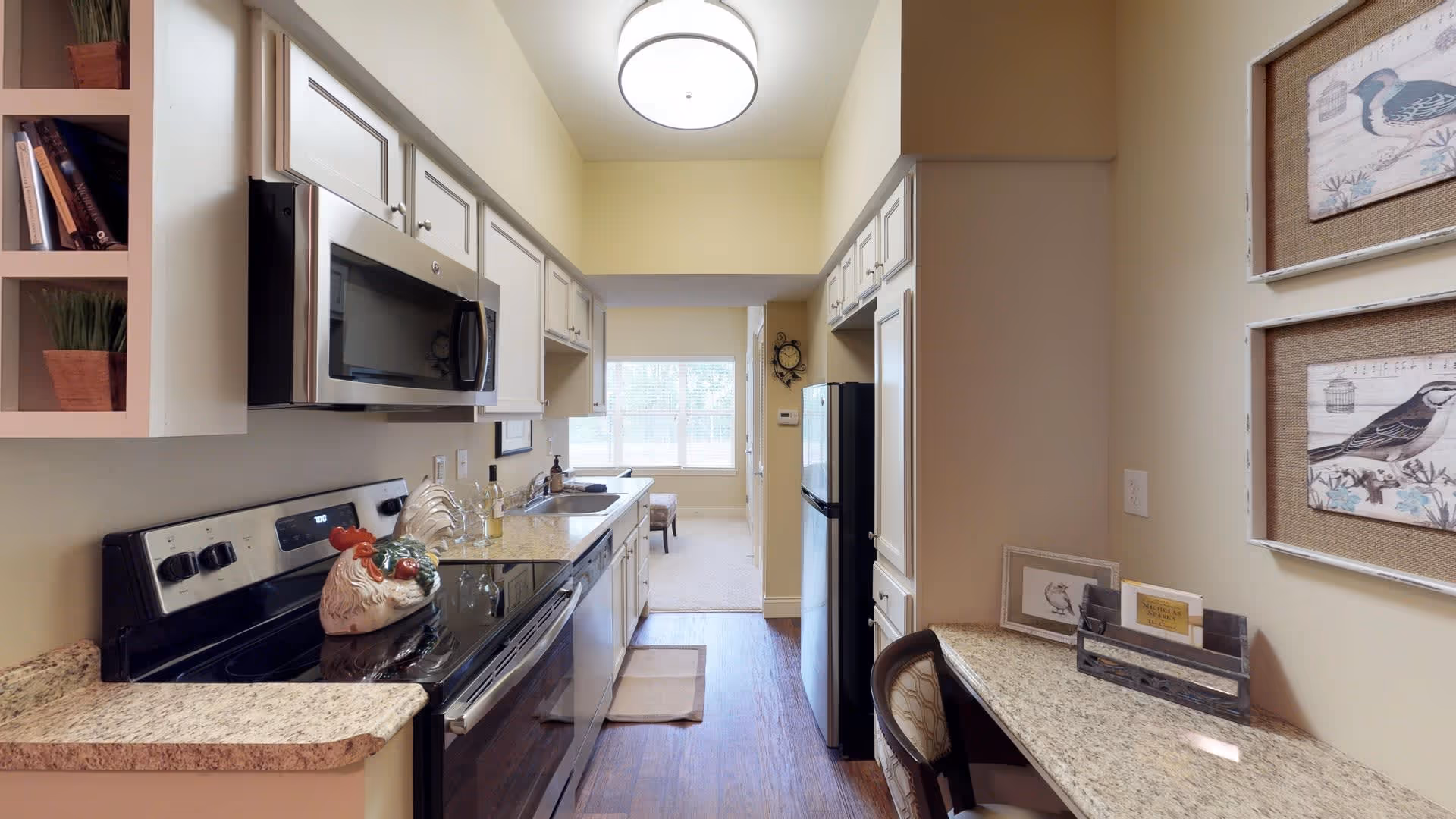 A narrow kitchen with granite countertops, white cabinets, a black stove with a decorative ceramic rooster on it, a microwave above the stove, a dishwasher, and a refrigerator. There is a small desk area with a chair and framed bird artwork on the wall. The kitchen leads to a bright room with a window and a chair visible in the background.