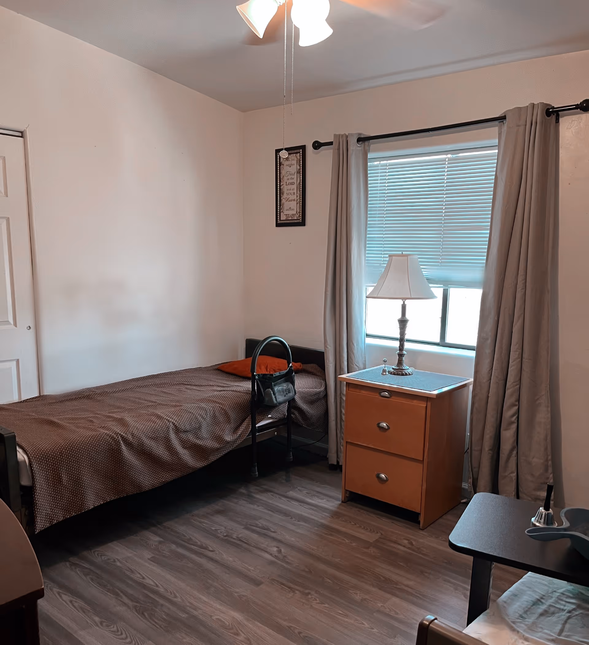 A simple bedroom with a single bed covered with a brown blanket and an orange pillow. A black handbag is hanging on a bed assist rail. Next to the bed is a wooden nightstand with two drawers, a table lamp, and a window with closed blinds and beige curtains. The room has wood flooring and white walls with a framed decorative sign hanging above the bed.