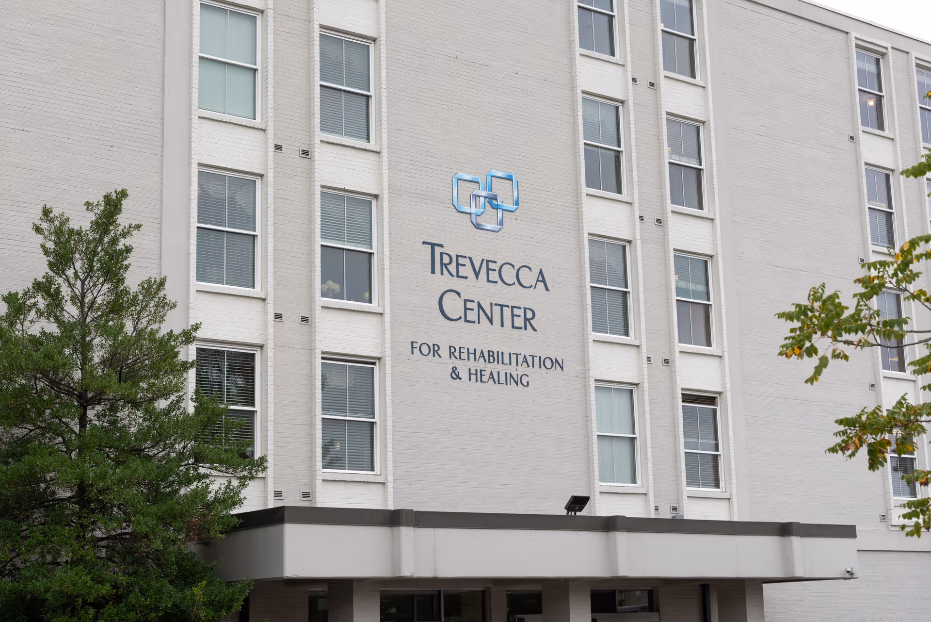 Exterior view of the Trevecca Center for Rehabilitation and Healing building, showing multiple windows and the facility's name and logo on the facade, with some trees partially visible in the foreground.
