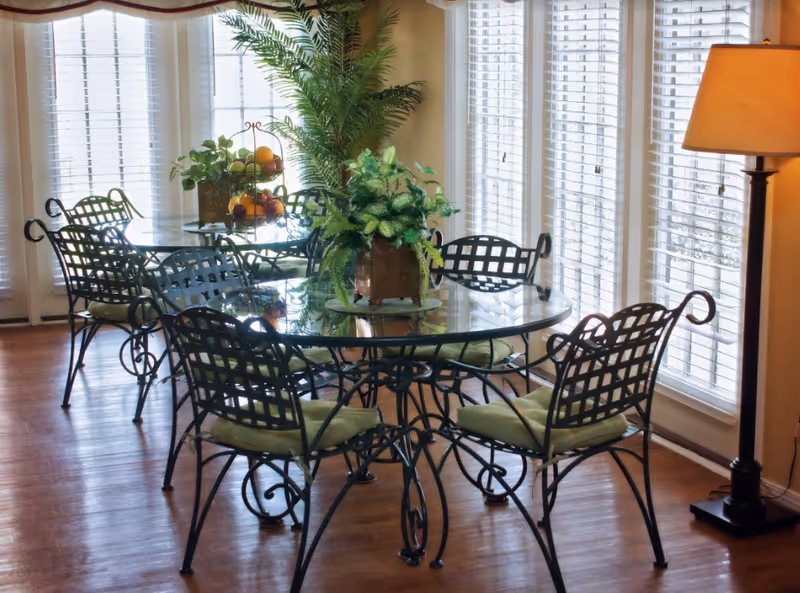 A bright dining area with two round glass tables surrounded by black metal chairs with green cushions. The room has large windows with white blinds allowing natural light to fill the space. There are green plants placed on the tables and a tall floor lamp in the corner.