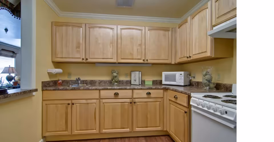 A kitchen with light wood cabinets, a brown countertop, a white microwave, a white electric stove with four burners, and a small sink. The walls are painted yellow and there is a pass-through window to an adjacent room with a lamp and some decor visible.