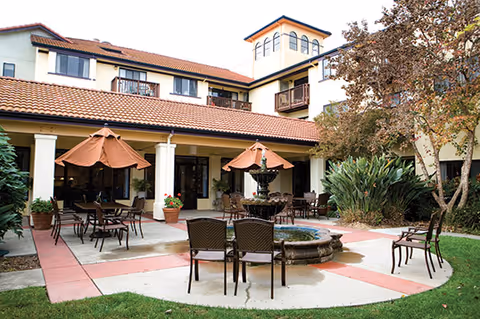 Outdoor patio area at Drake Terrace featuring multiple tables with umbrellas, chairs, a central water fountain, and surrounding greenery including trees and shrubs. The building with a tiled roof and balconies is visible in the background.