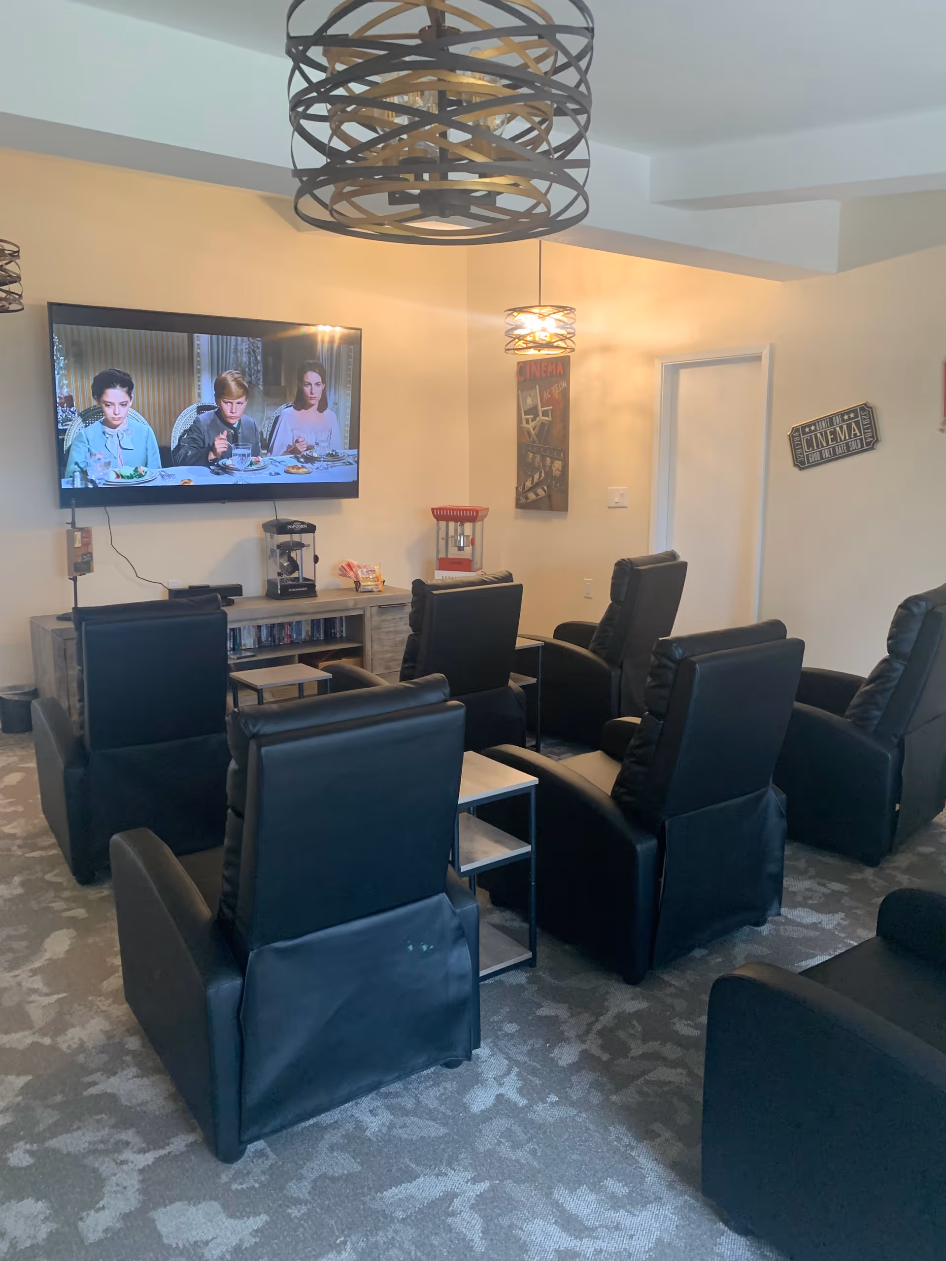 A small theater room with six black recliner chairs arranged in two rows facing a wall-mounted flat screen TV showing a movie scene with three people eating at a table. The room has beige walls, carpeted floor, two modern pendant lights, a popcorn machine on a wooden cabinet below the TV, and cinema-themed wall decorations.