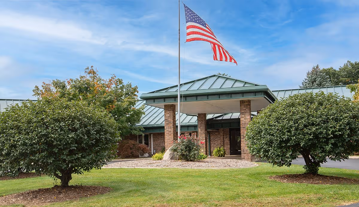 Exterior view of a senior living facility with a green metal roof and brick pillars supporting a covered entrance. An American flag is flying on a flagpole in front of the building, surrounded by neatly trimmed bushes and a well-maintained lawn under a partly cloudy sky.